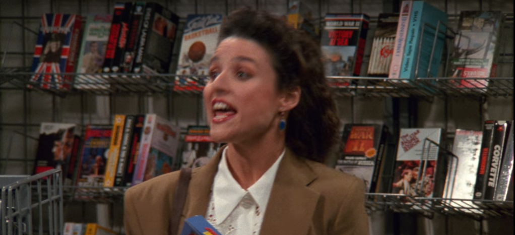 A screenshot from Seinfeld of Elaine talking in front of a row of VHS tapes in a video store.