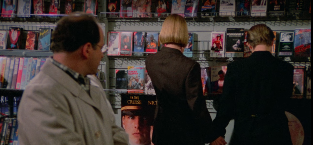 A screenshot from Seinfeld of George looking at Susan and her girlfriend browse a wall of VHS tapes in a rental store.